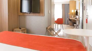 Habitación de hotel con cama, televisor montado en la pared, escritorio con silla y baño visible a través de una puerta de cristal.