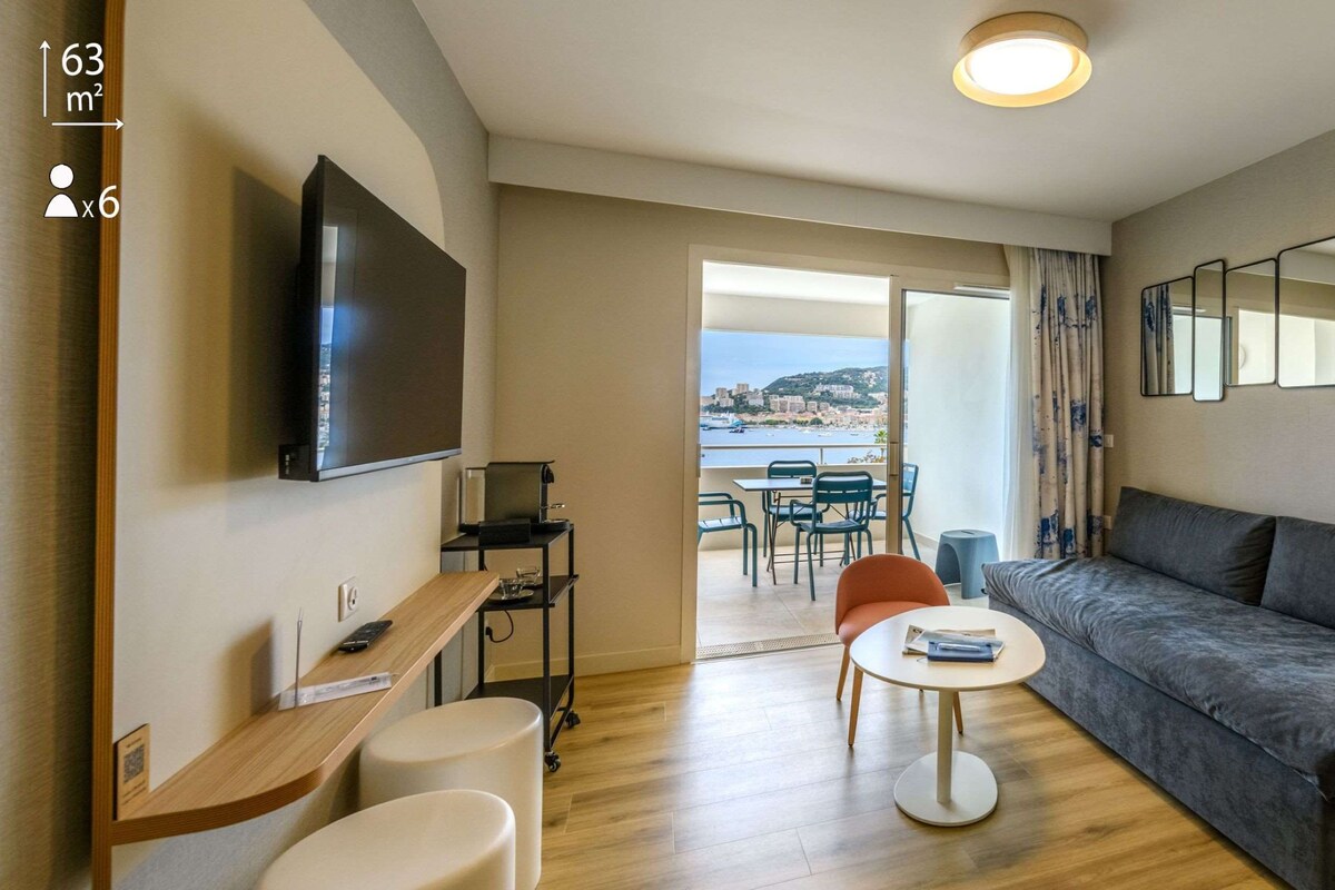 Una sala moderna con un sofá, un televisor, una mesita y un balcón con vistas al mar.