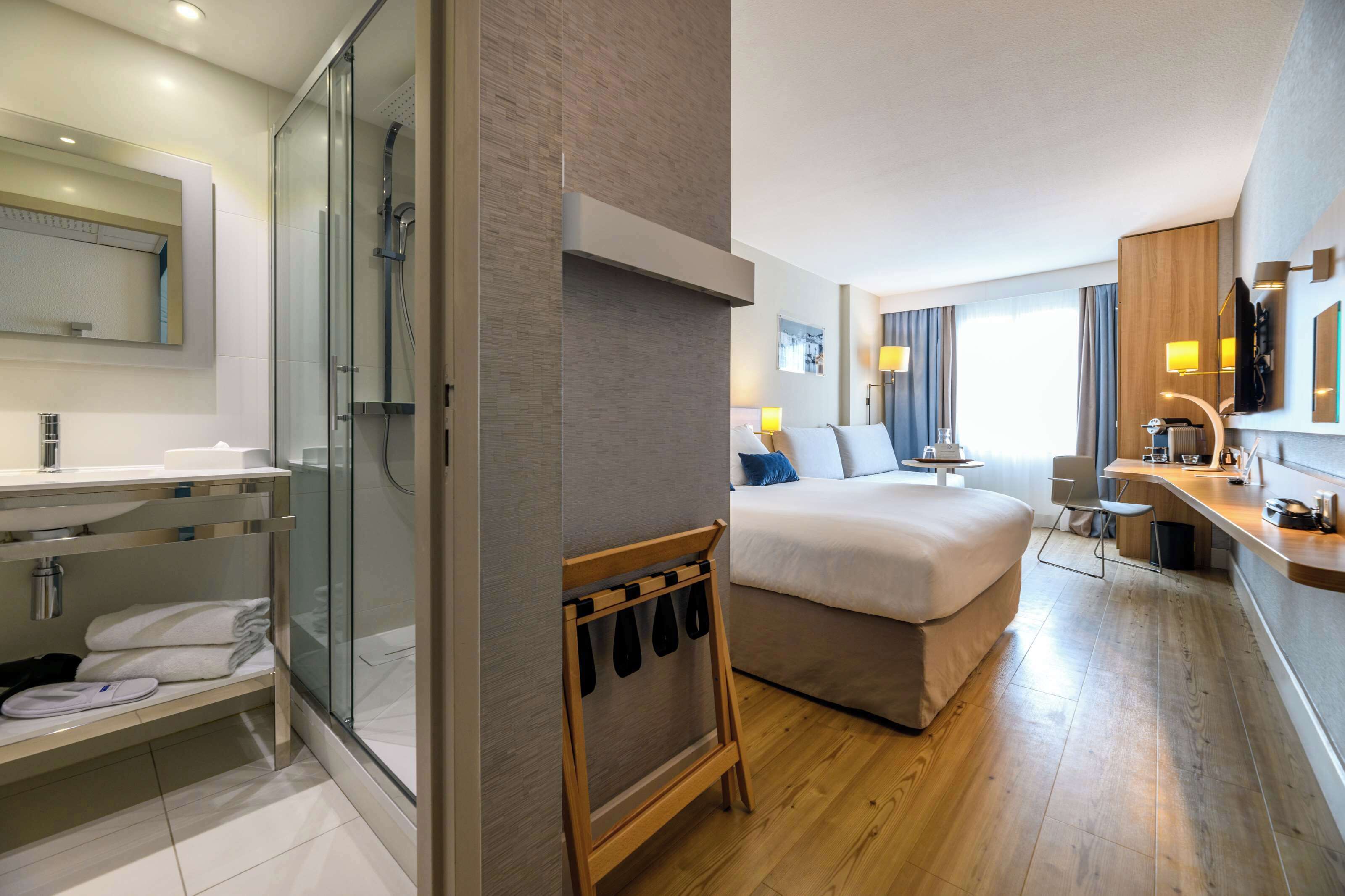 A modern hotel room with a shower, a bed, a desk, and a bathroom with a sink and mirror.