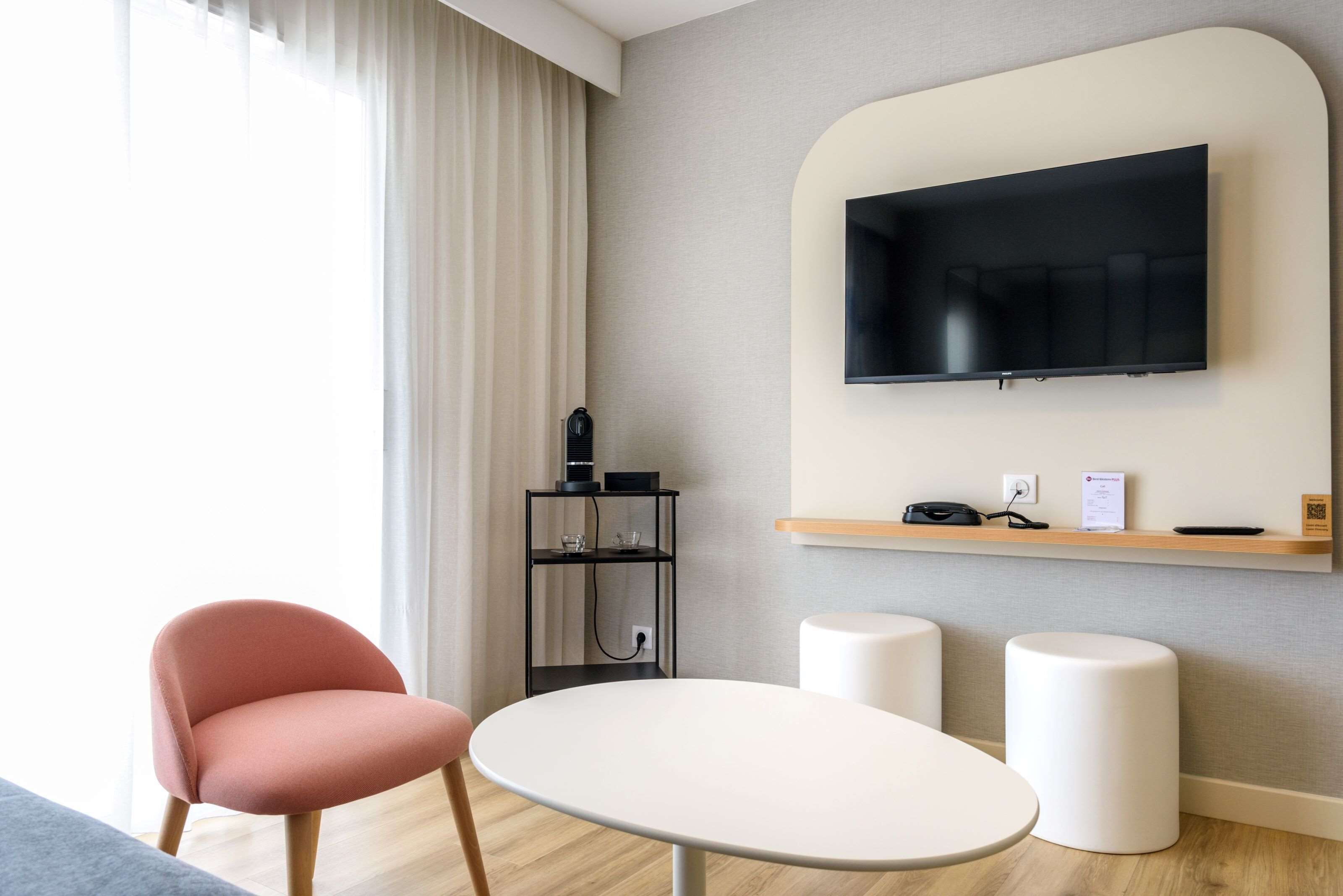 Een moderne hotelkamer met een flatscreen-tv aan de muur, een klein houten plankje met een telefoon en een ingelijst certificaat, een ronde witte tafel, twee witte krukken, een roze stoel, en een zwart plank met glazen en een koffiezetapparaat.