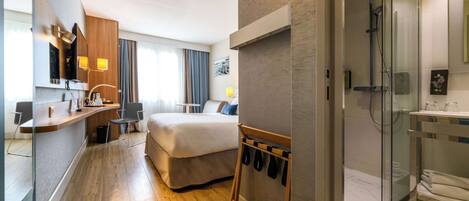 A modern hotel room with a bed, a desk with a chair, a shower area, and a coat rack.