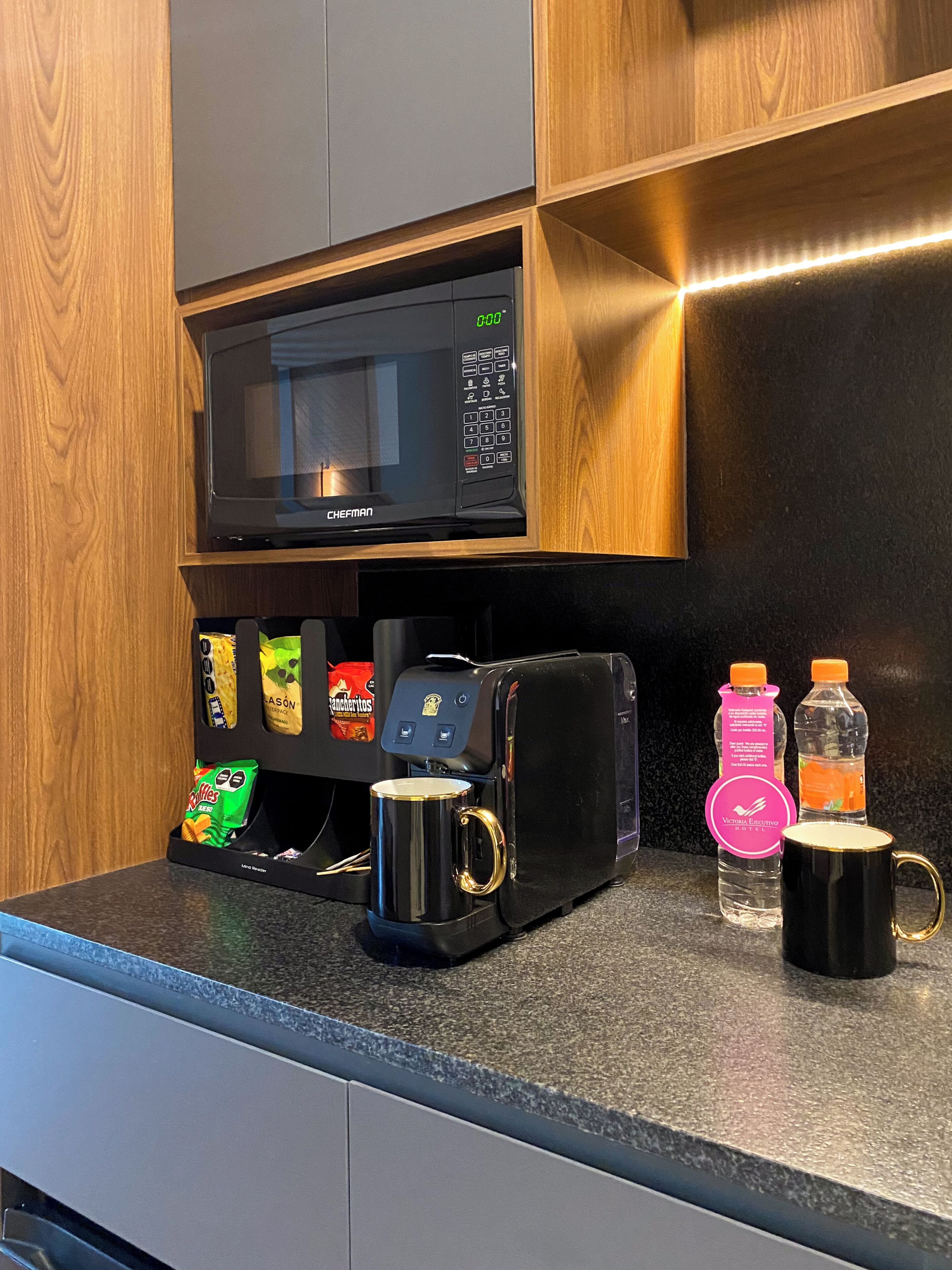 A kitchen with a microwave, coffee machine, snacks, and two mugs on the counter.