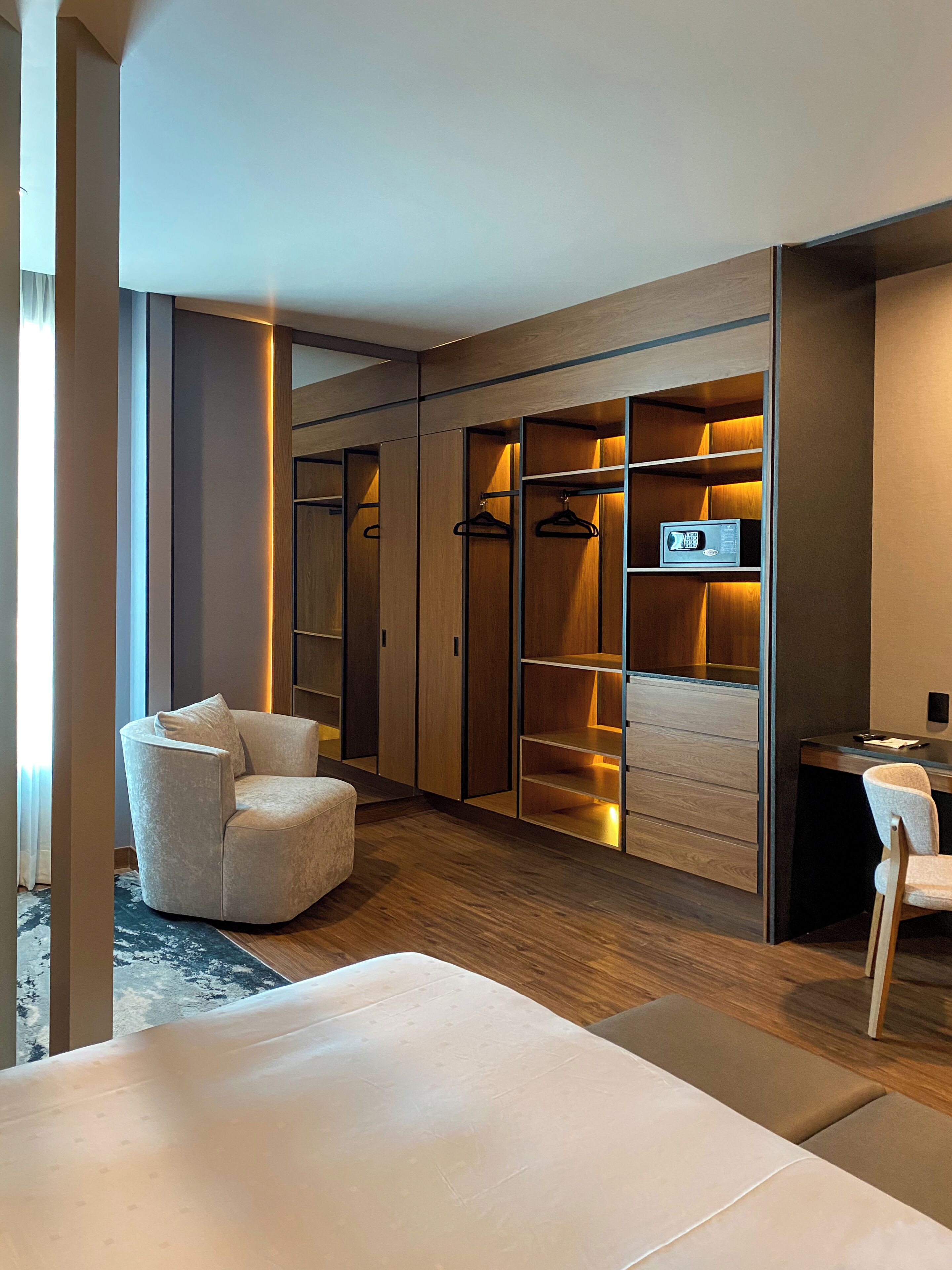 Una habitación de hotel moderna con zona de estar, escritorio y vestidor con espacio para colgar y estantes.