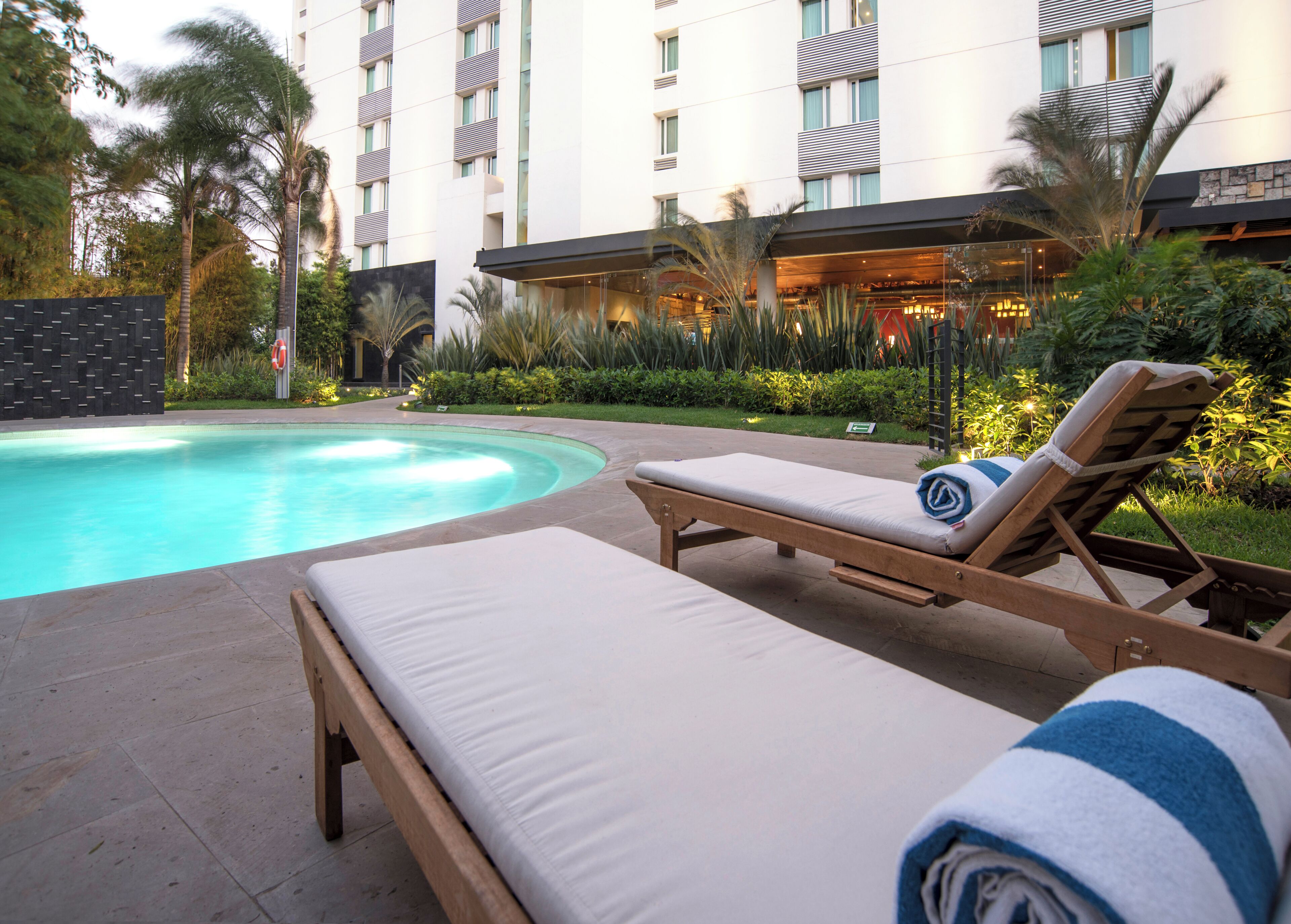 Zona de piscina con sillones de descanso y una piscina, rodeada de vegetación y un edificio de varios pisos.