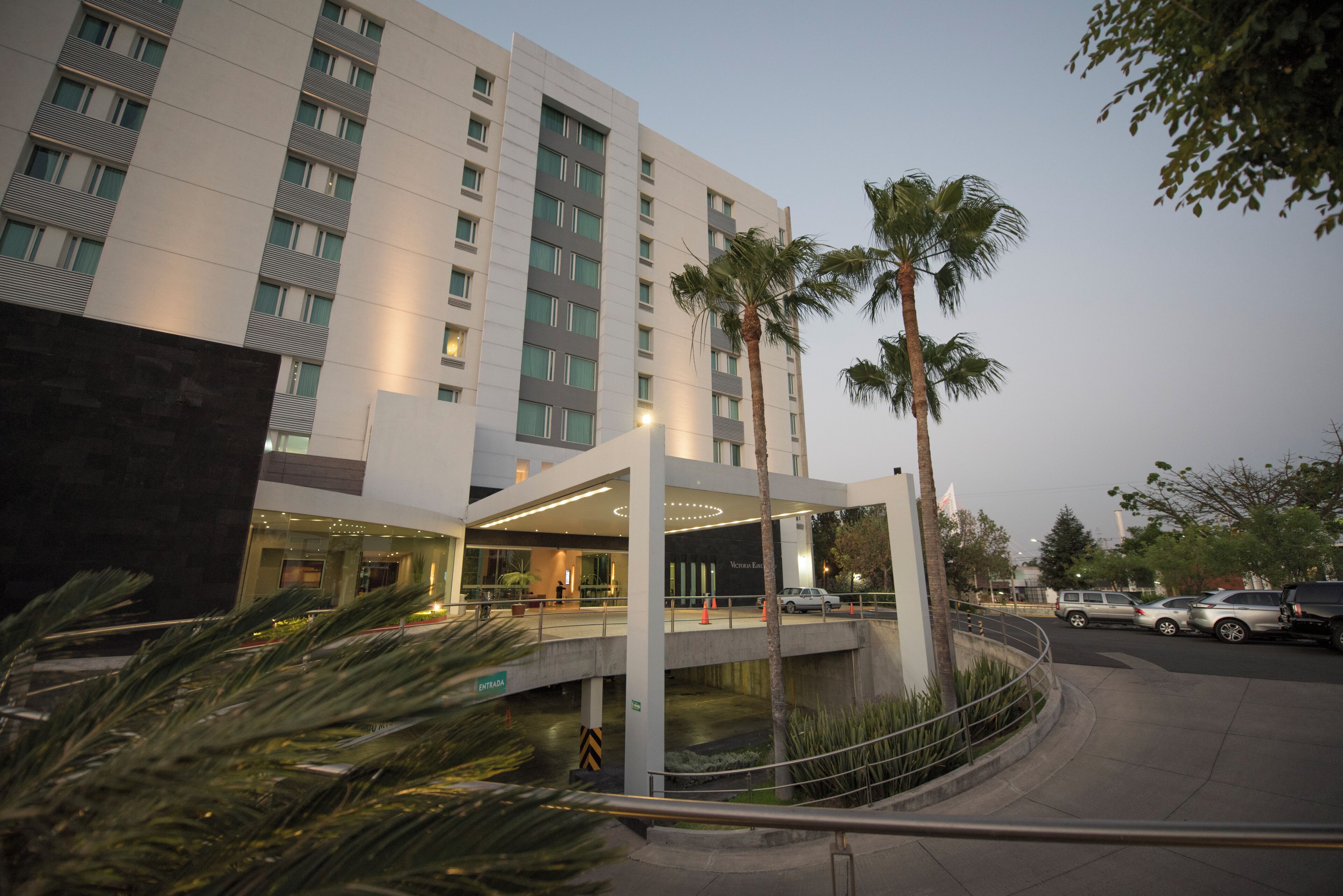 A modern hotel with a covered entrance, palm trees, and a parking area.