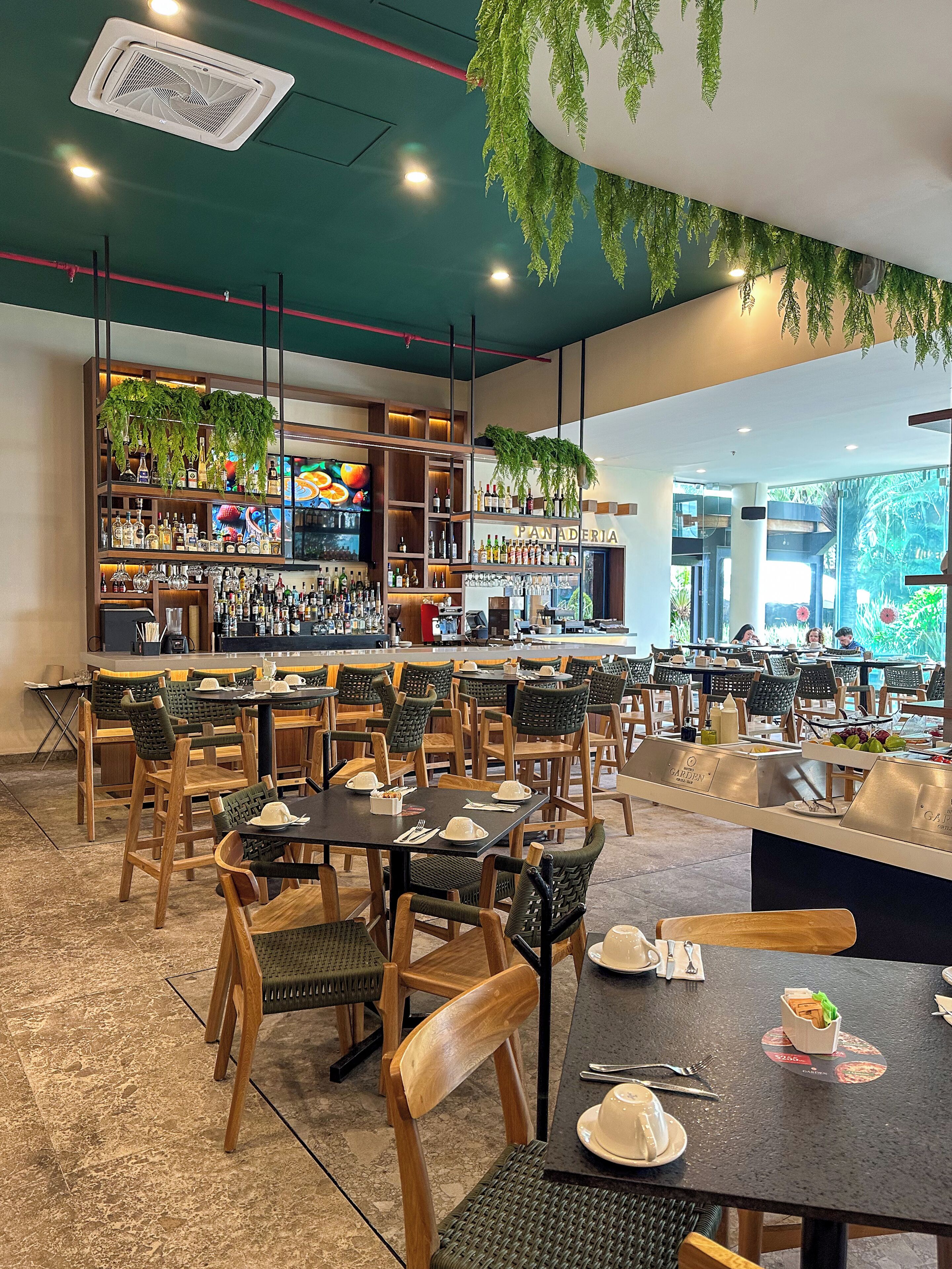 Interior de restaurante con varias mesas, sillas, una zona de bar con estantes llenos de botellas y plantas colgantes.
