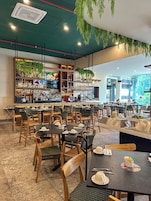 Interior de restaurante con varias mesas, sillas, una zona de bar con estantes llenos de botellas y plantas colgantes.