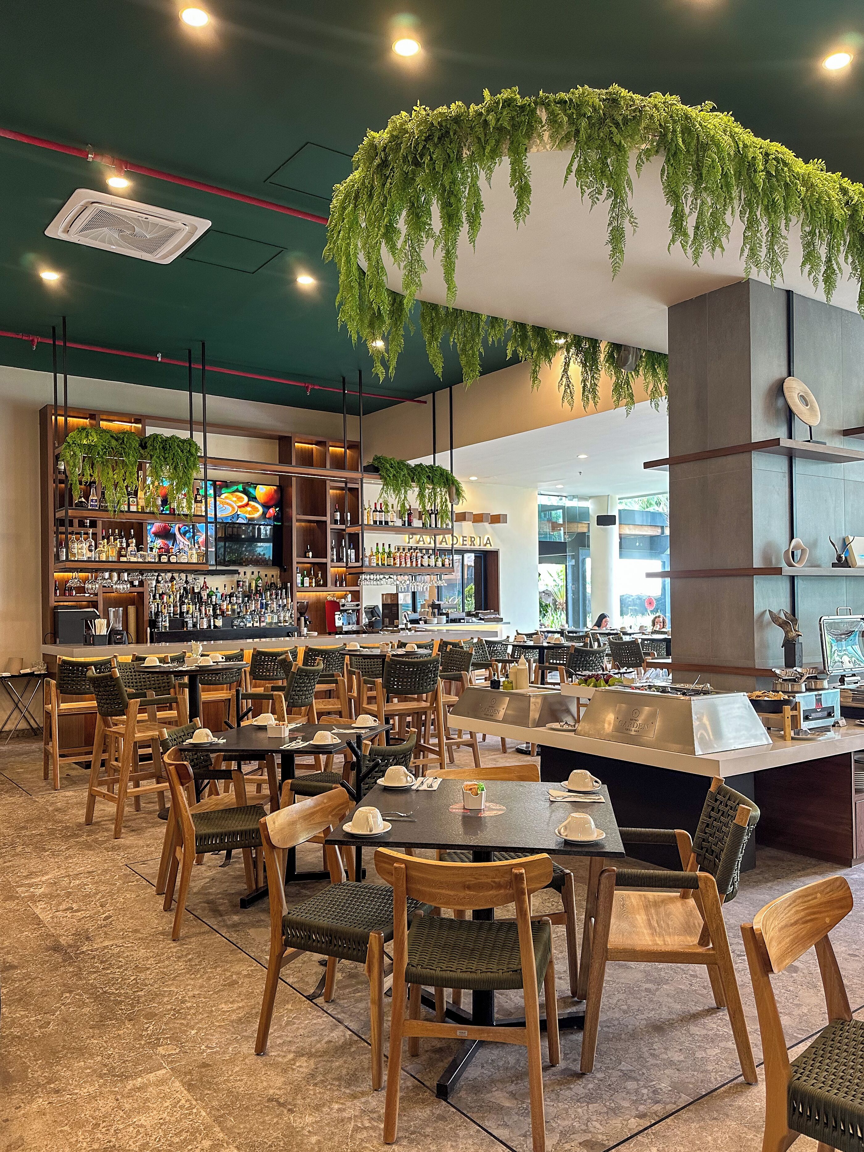 A restaurant with multiple dining tables and chairs, a bar area with shelves of bottles, and hanging greenery.