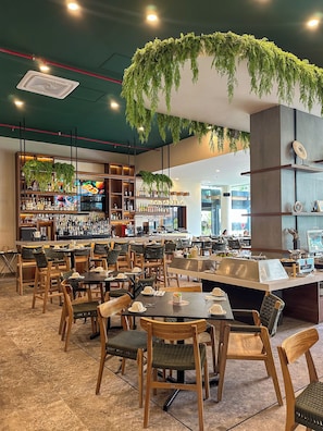 A restaurant with multiple dining tables and chairs, a bar area with shelves of bottles, and hanging greenery.