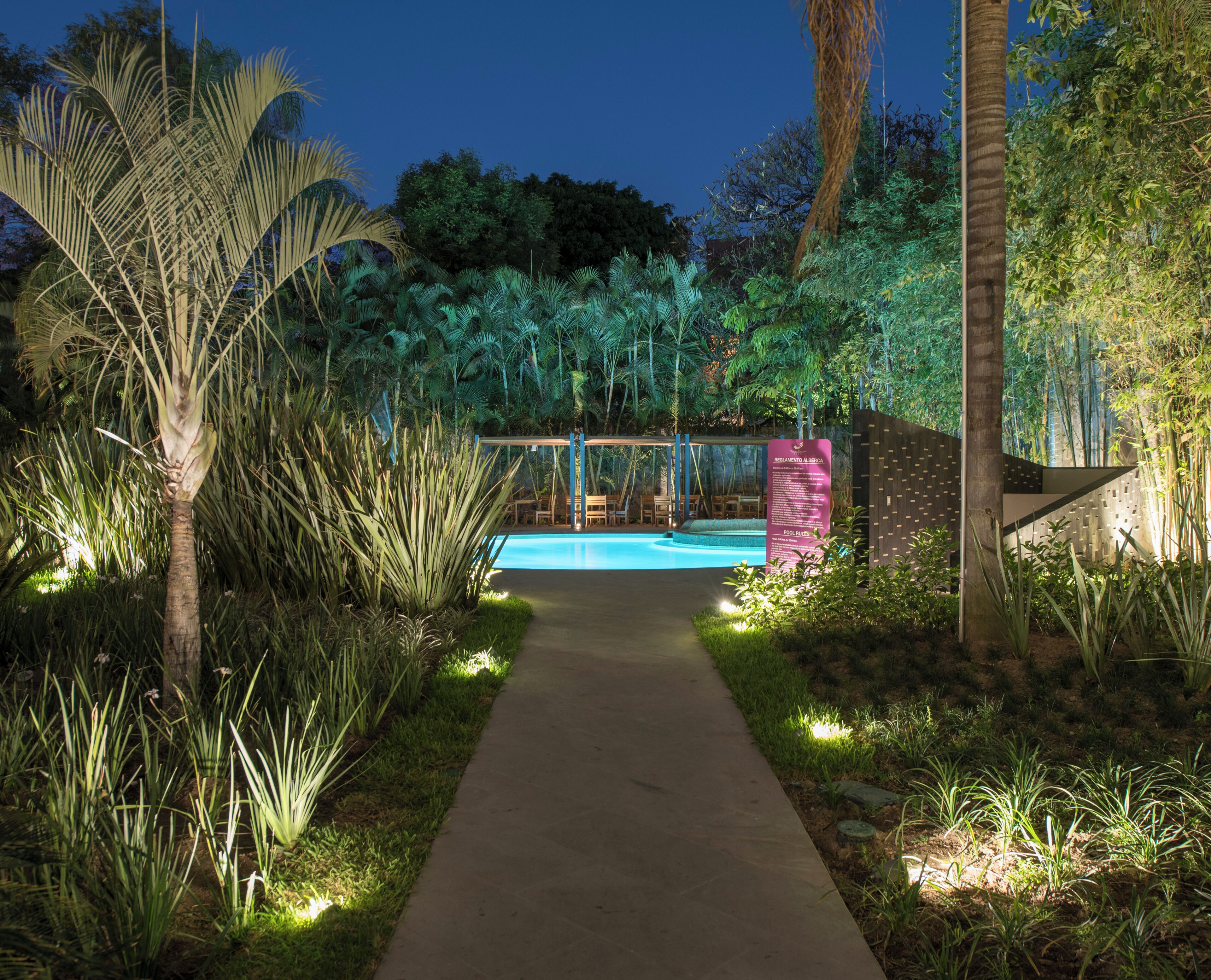 Un sendero conduce a una piscina rodeada de exuberante vegetación y una valla de madera.