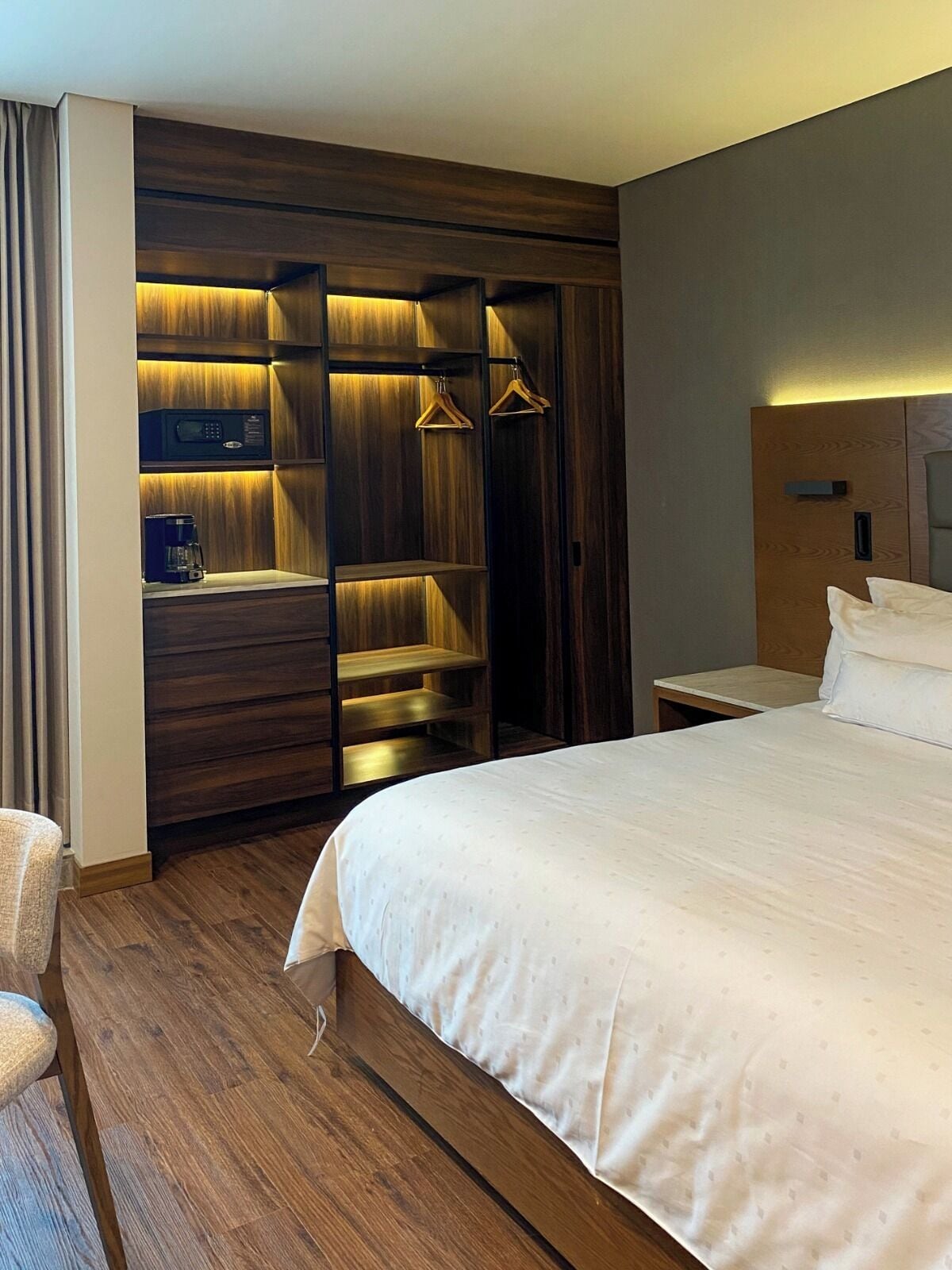 A hotel room with a large bed, a wooden wardrobe with built-in lighting, a bedside table, and a chair.