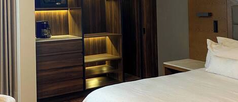 Habitación de hotel con una cama grande, un armario de madera con iluminación integrada, una mesita de noche y una silla.