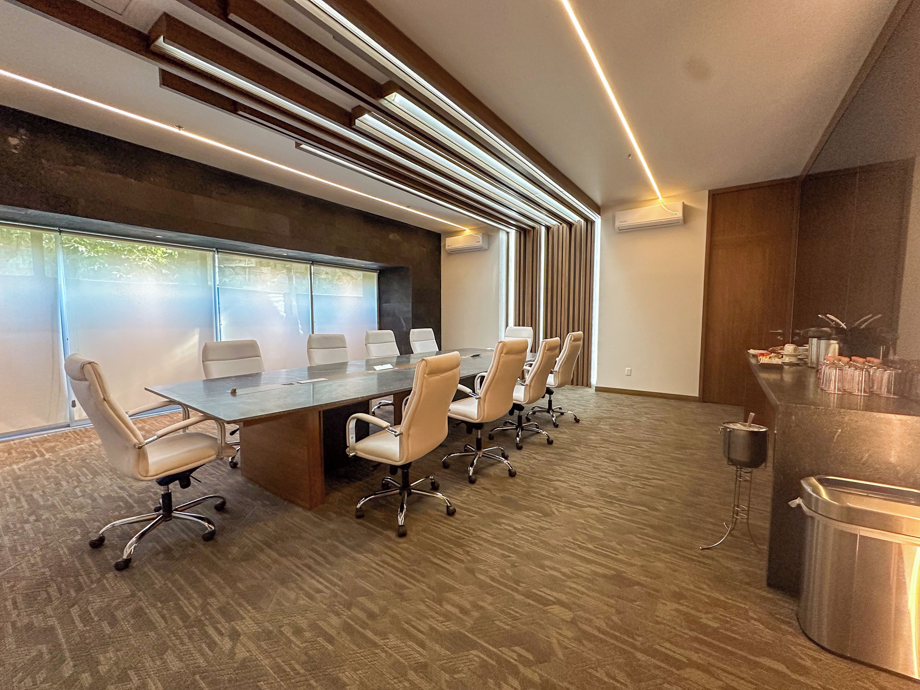 A conference room with a long table, multiple chairs, a modern ceiling design, and a large window with blinds.