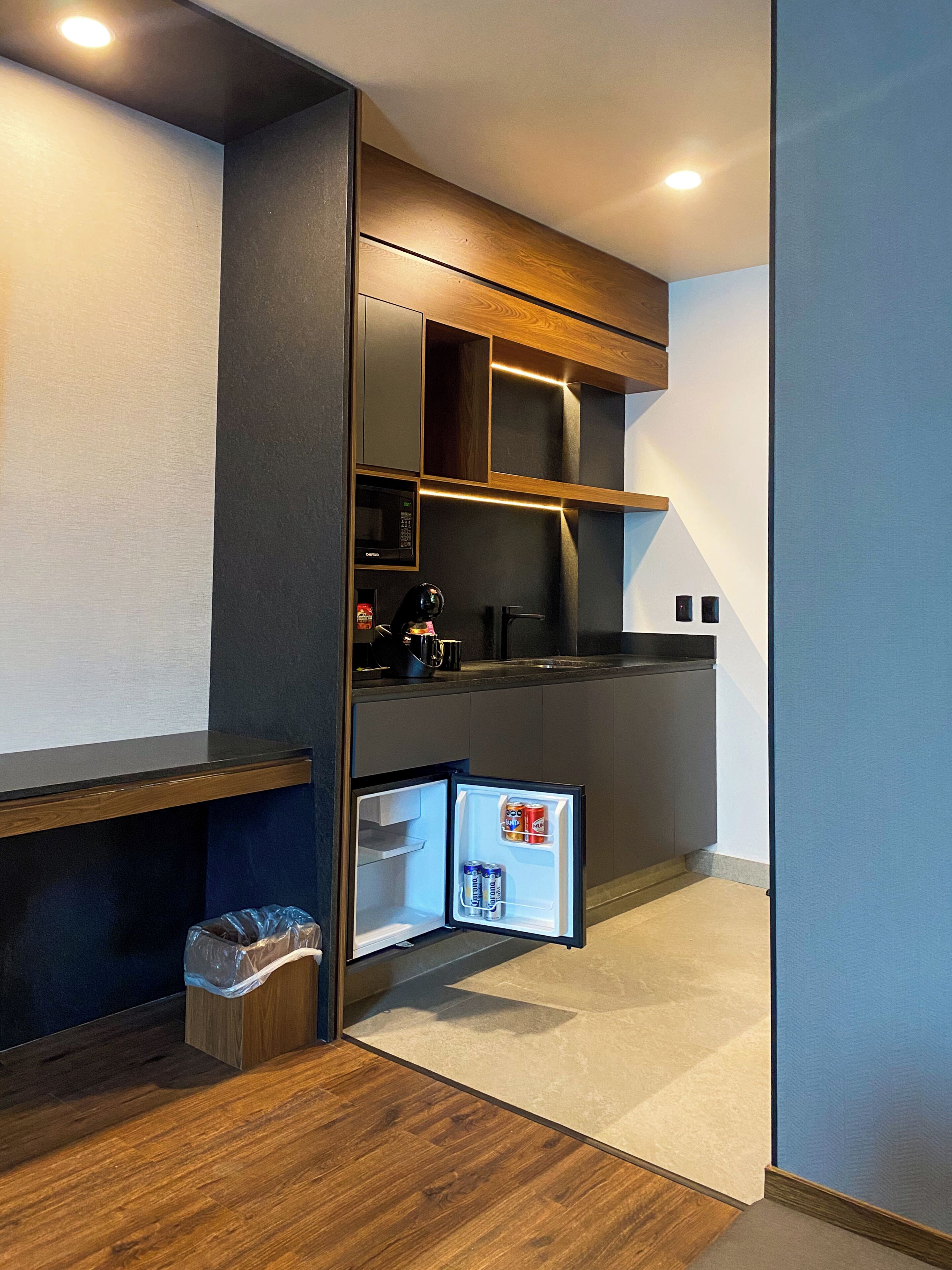 A compact kitchen with a built-in microwave, a small refrigerator, and a trash bin.