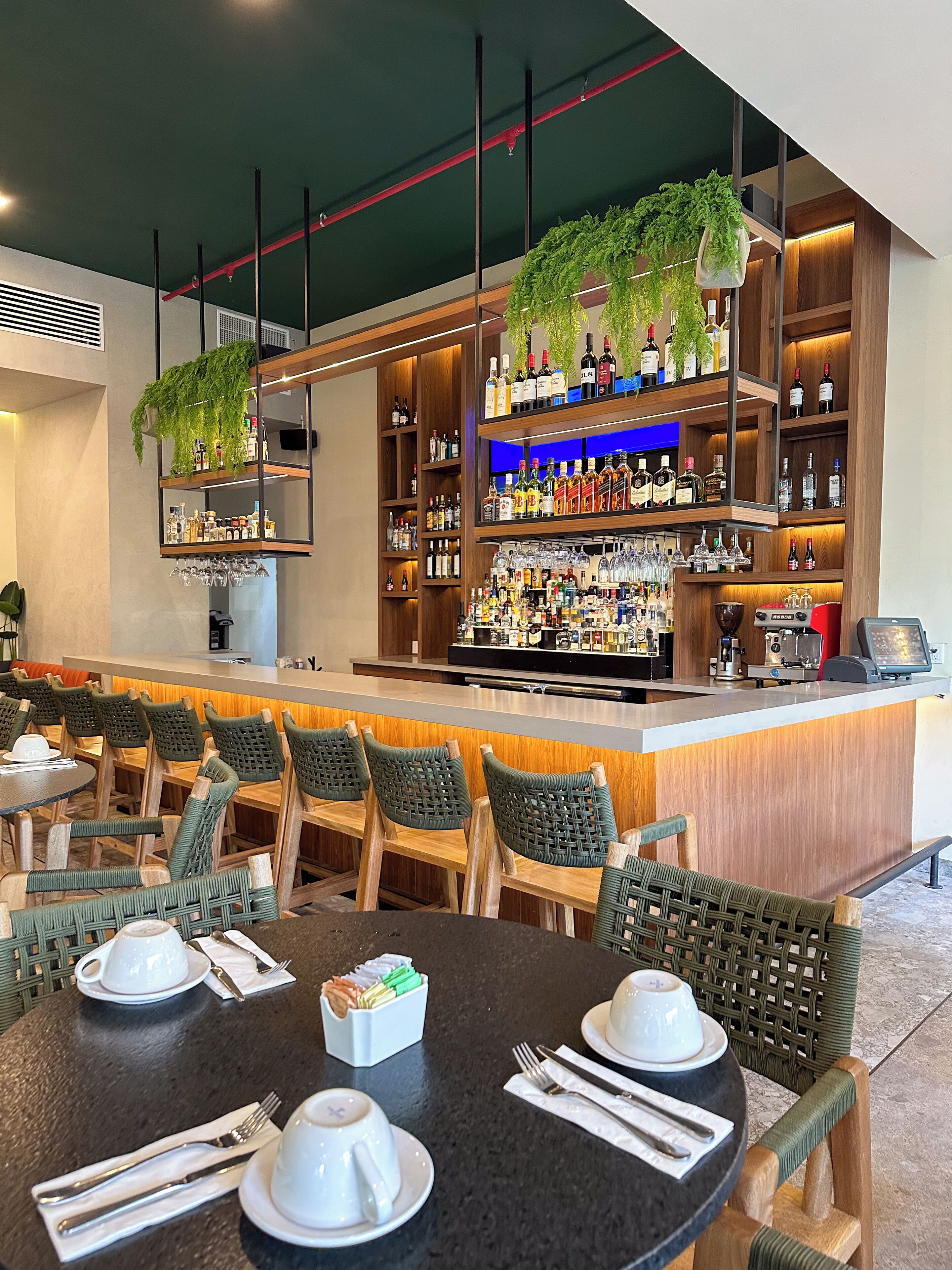 Una zona de bar con varias estanterías llenas de botellas de licores y vasos, sillas de madera y una barra con una máquina de café.