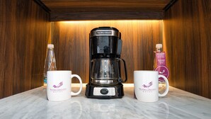 Une machine à café posée sur un comptoir en marbre, avec deux tasses blanches ornées d’un logo, une bouteille d’eau et une boîte de thé.
