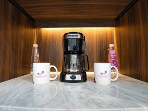 A coffee maker on a marble countertop with two white mugs with a logo, a bottle of water, and a box of tea. - Hotel Victoria Ejecutivo (Guadalajara)