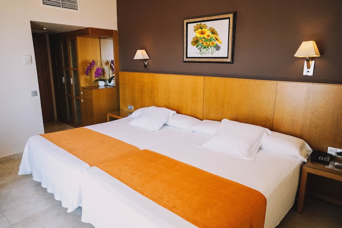 A hotel room with a large bed, a wooden headboard, two bedside lamps, a framed floral artwork on the wall, and a small table with a phone and a lamp.