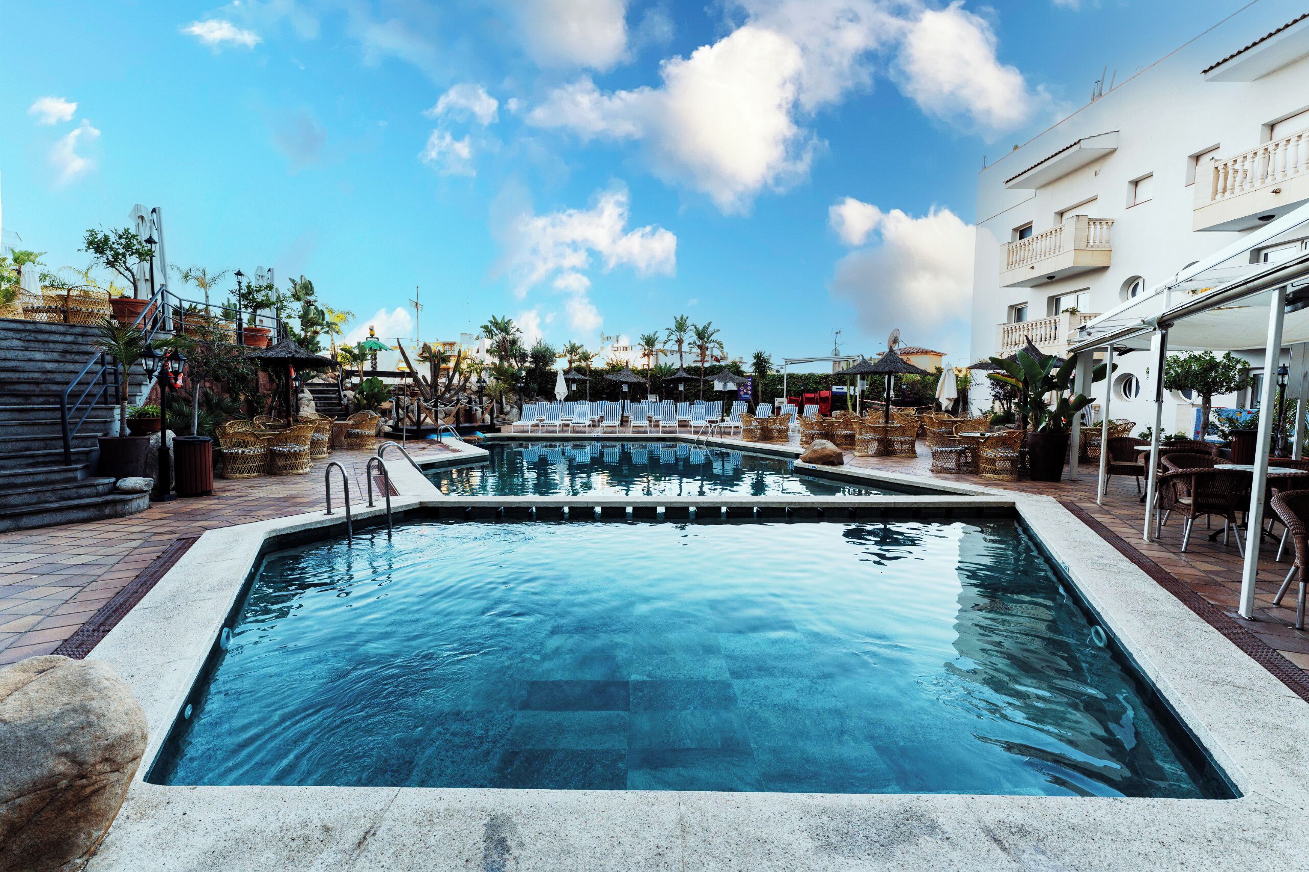 Una piscina con sillas de descanso y sombrillas, rodeada de palmeras y un edificio con balcones.