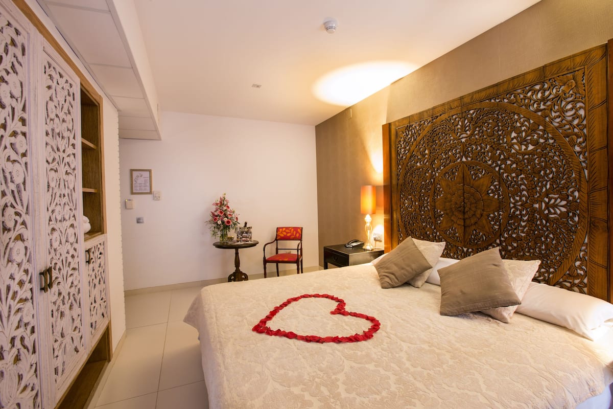 A hotel room with a bed, a decorative headboard, a nightstand with a lamp, a chair, and a small table with a vase of flowers.