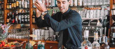 Un cantinero preparando una bebida en la barra, con estantes llenos de botellas y vasos al fondo.