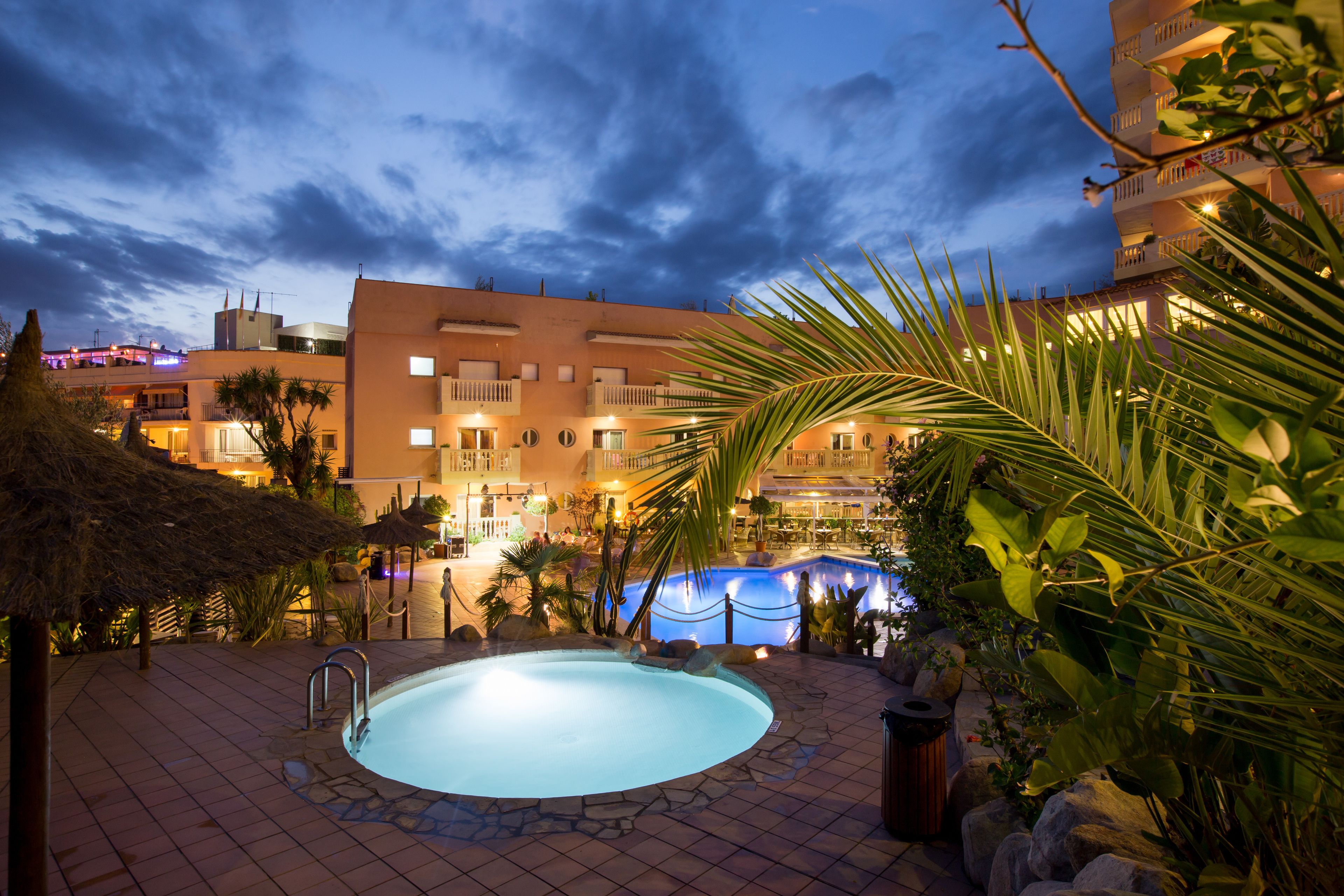 Zona de piscina con solárium de baldosas de piedra, rodeada de palmeras y un edificio con varios balcones.