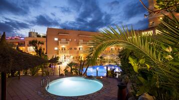 Zona de piscina con solárium de baldosas de piedra, rodeada de palmeras y un edificio con varios balcones.