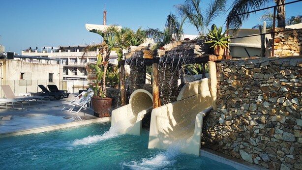A pool with a waterfall feature, lounge chairs, and a stone wall. - Alba Seleqtta (Lloret de Mar)