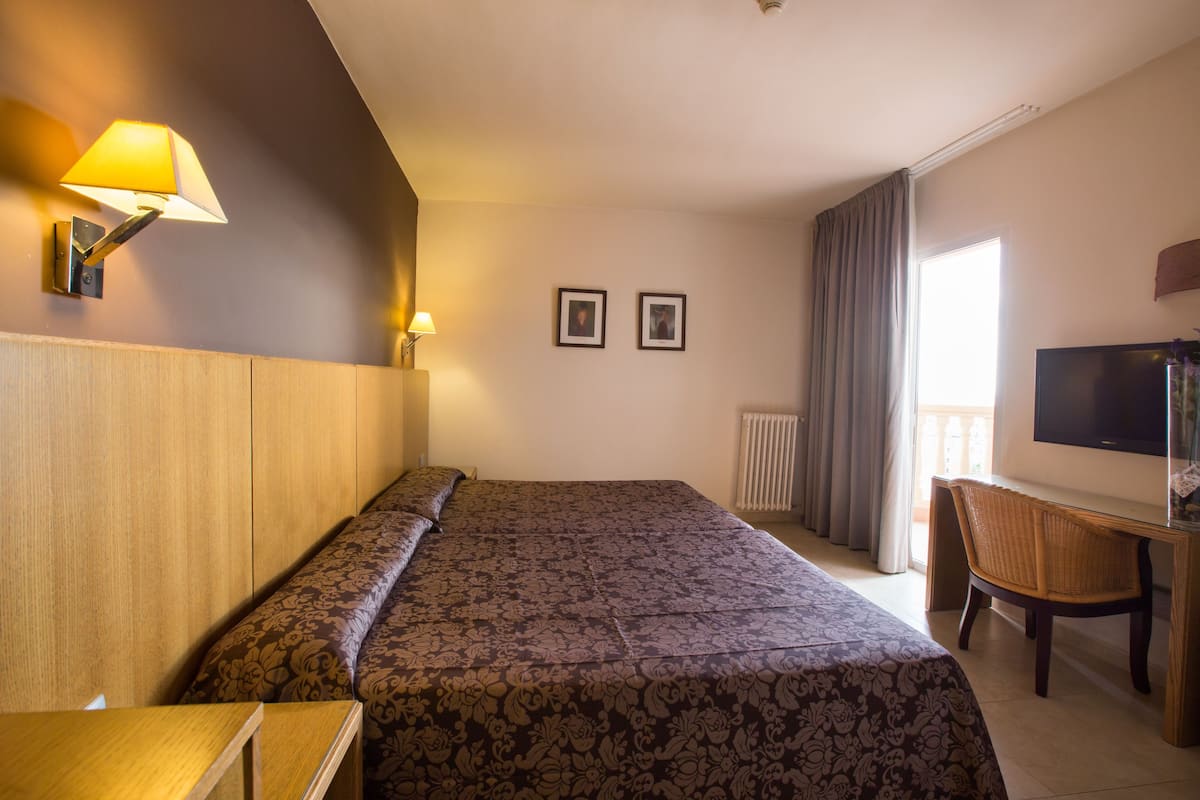 A hotel room with a bed, a desk, a chair, a TV, and two framed pictures on the wall.