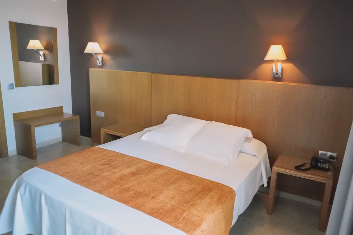 A hotel room with a large bed, a wooden headboard, two bedside tables with lamps, a small wooden bench, and a mirror on the wall.