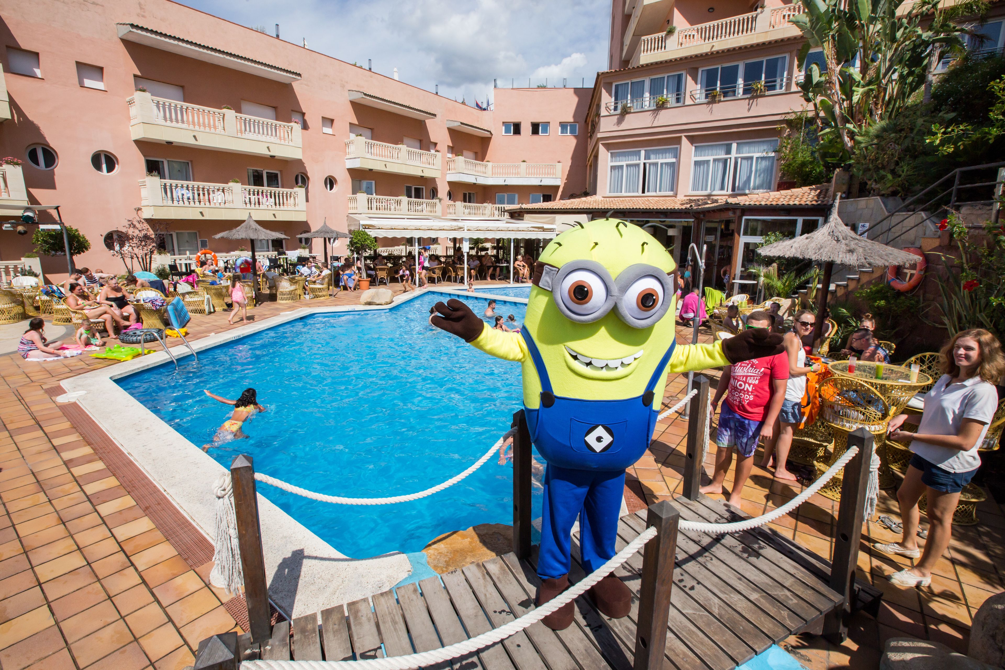 Zona de piscina con un personaje mascota, sillas de descanso y un edificio con balcones.
