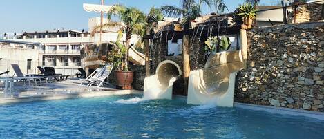 A pool with a water slide, lounge chairs, and a stone wall.