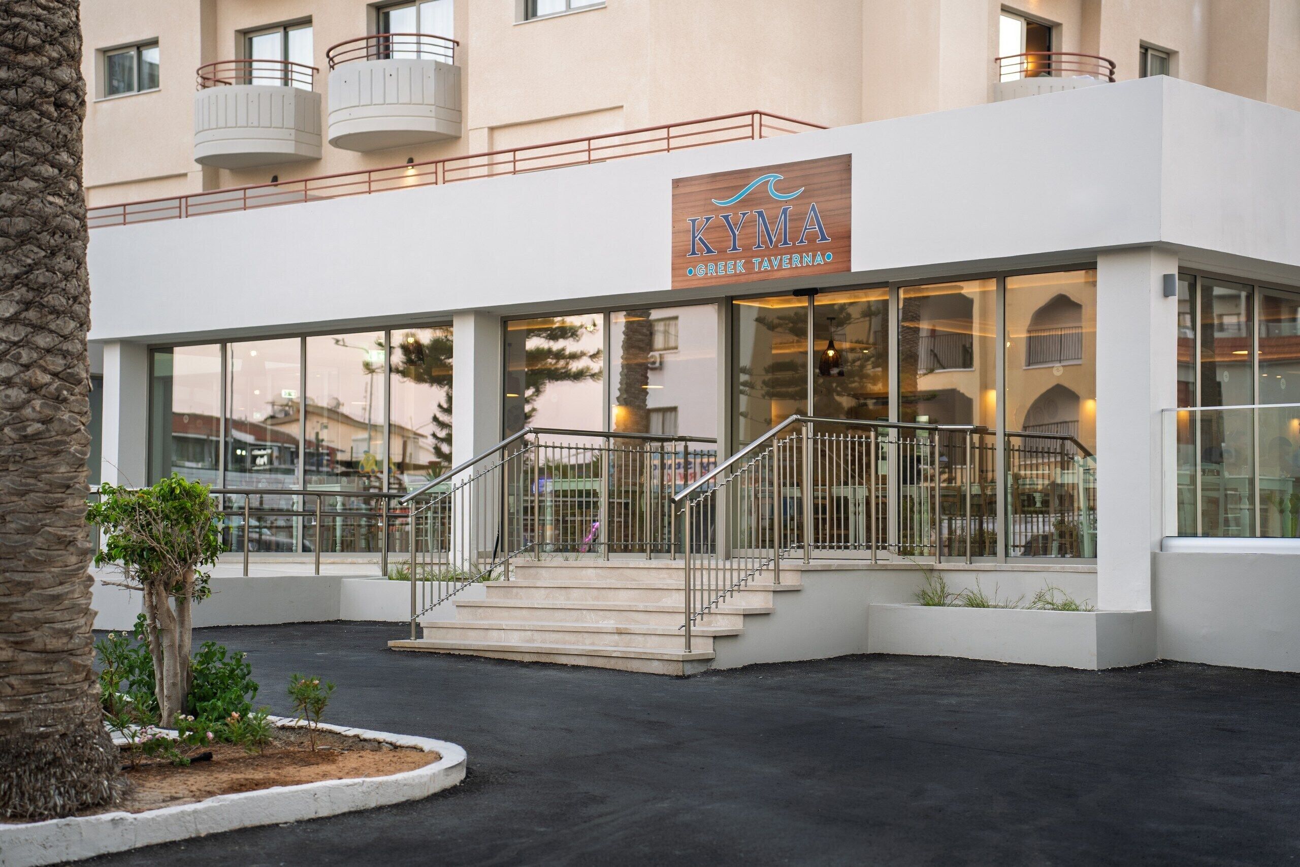 A building with a sign reading 'KYMA GREEK TAVERN' and a staircase leading to an entrance.