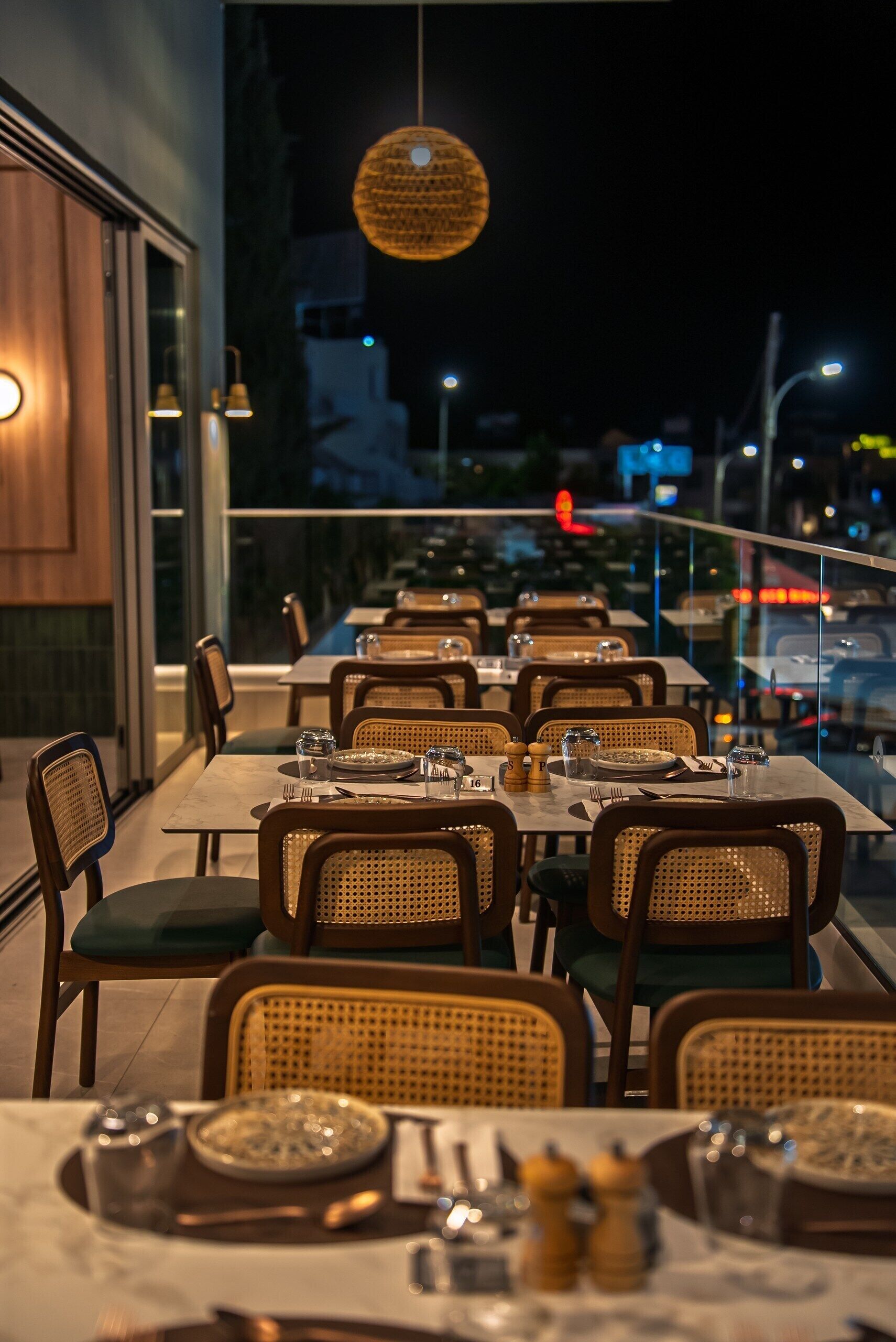 Un área de comedor al aire libre con mesas dispuestas para dos, sillas de mimbre y una lámpara colgante.