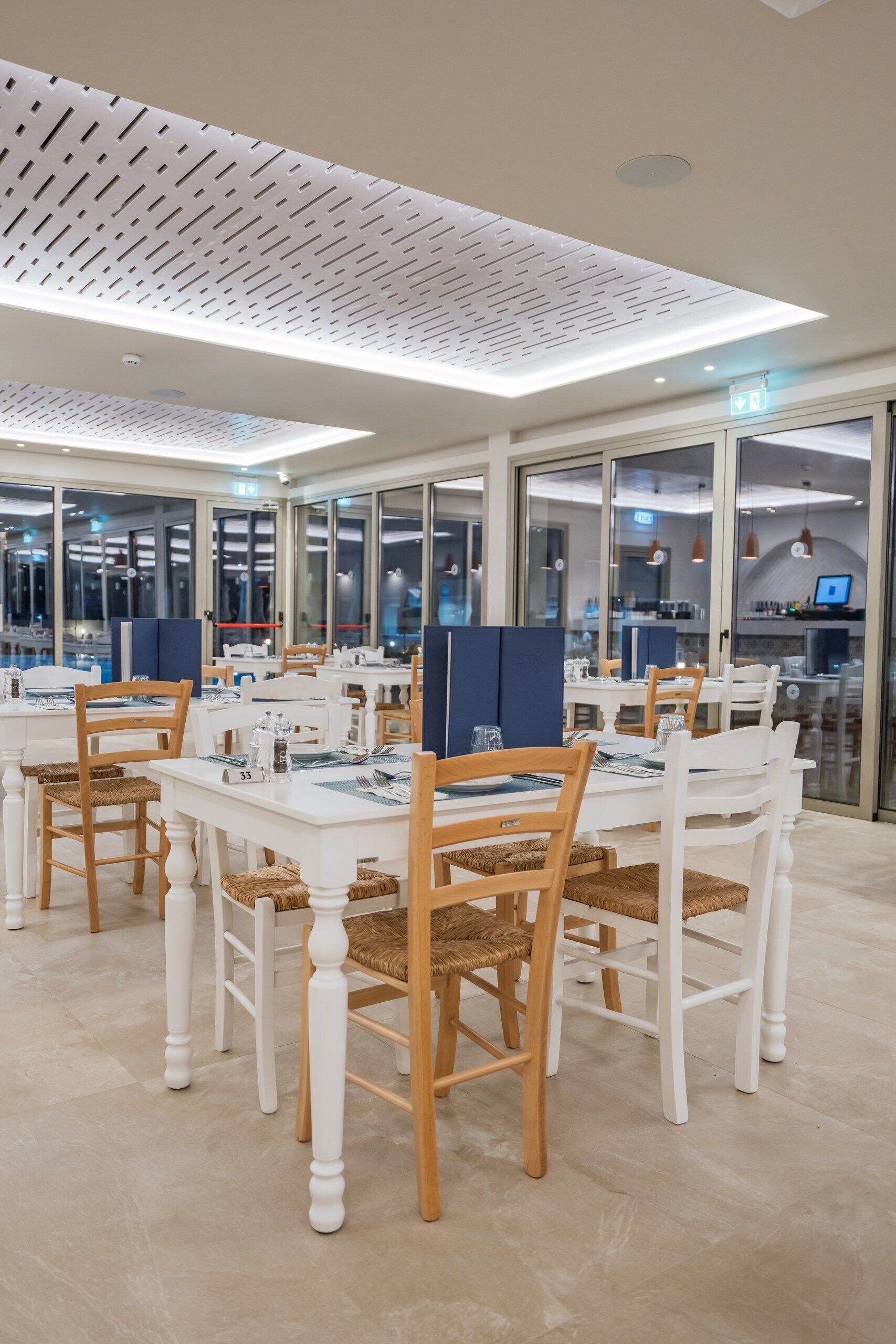 Un comedor moderno con mesas y sillas blancas, divisiones de vidrio y un techo con diseño perforado.