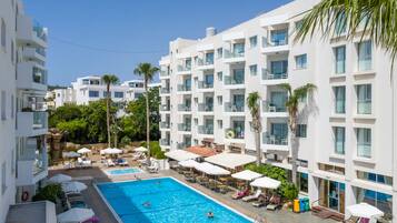 A modern hotel with a swimming pool surrounded by sun loungers and umbrellas, and multiple balconies with clear views.