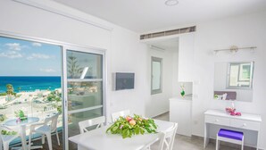Habitación moderna con balcón y vista a la playa, un escritorio blanco y un área de comedor con sillas.