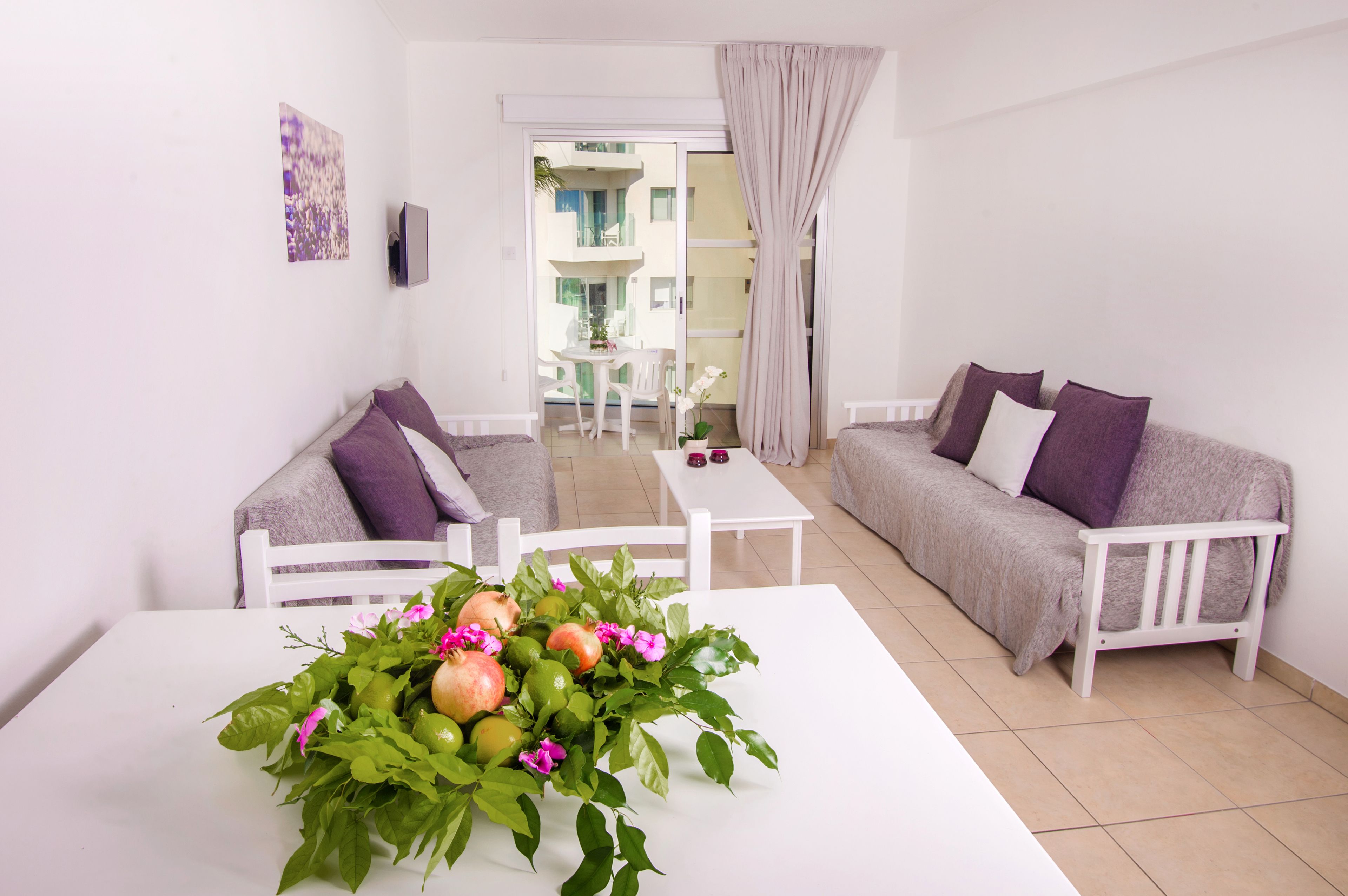 Ein modernes Wohnzimmer mit einem weißen Sofa, einem weißen Tisch mit einer Frucht- und Blumenkomposition und Blick auf den Balkon.