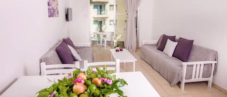 A modern living room with a white sofa, a white table with a fruit and flower arrangement, and a view of the outside balcony.