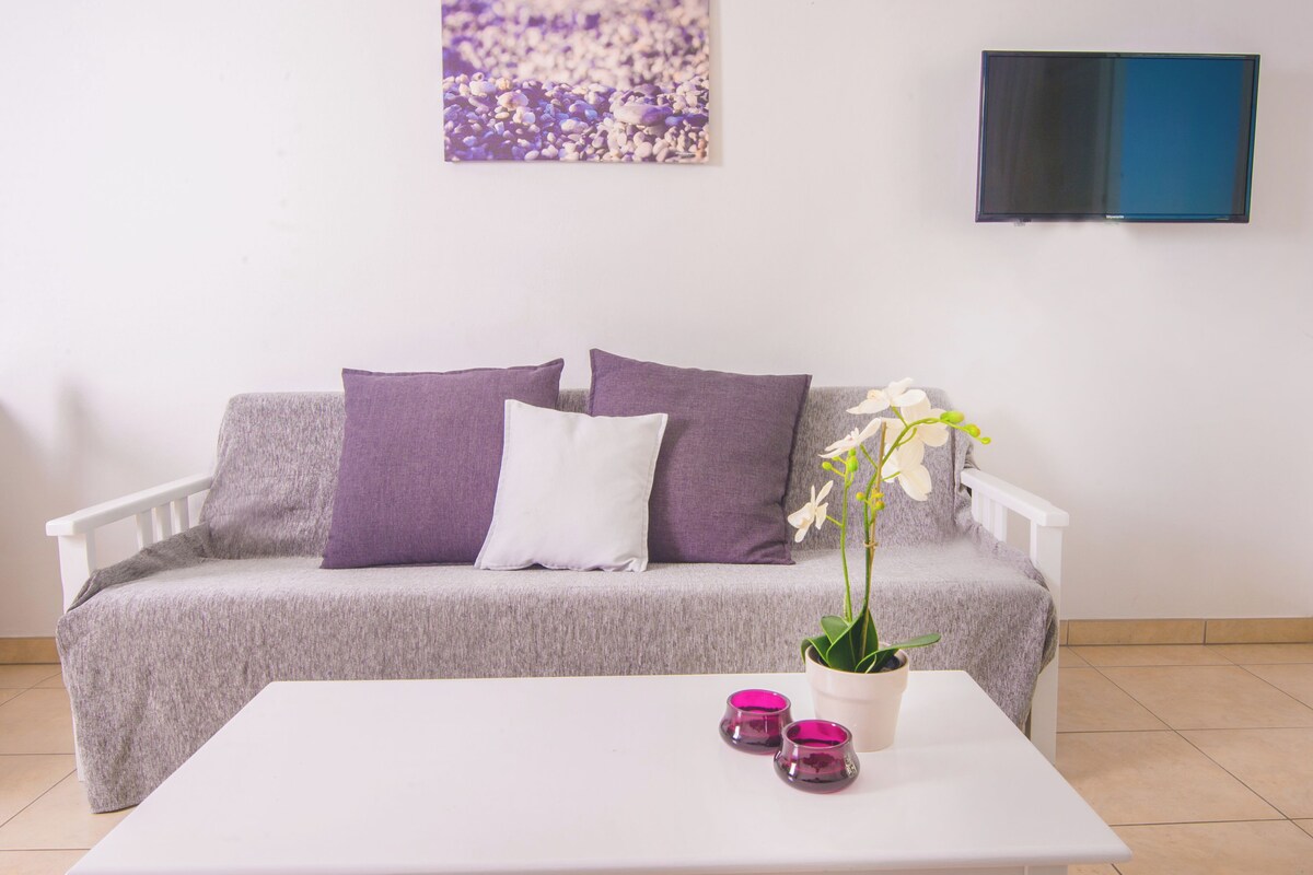 A modern living room with a white sofa, purple cushions, a white coffee table, and a potted orchid.