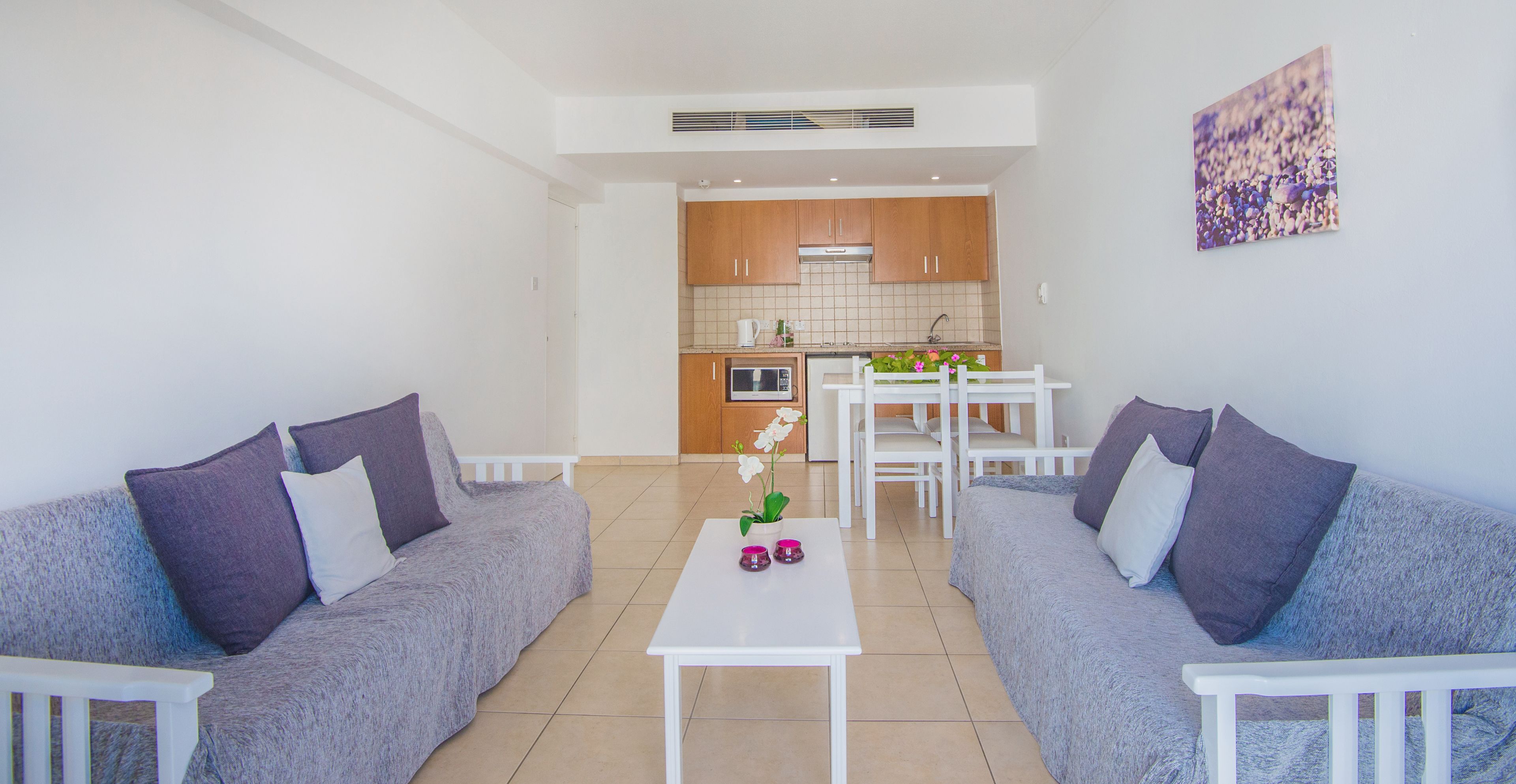 Una sala moderna con dos sofás grises, una mesa de centro blanca con objetos decorativos, y vistas hacia la zona de la cocina con armarios de madera y una mesa de comedor.