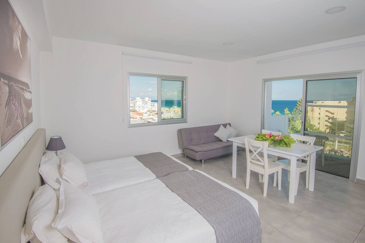 A modern hotel room with a bed, a sofa, a dining table with chairs, and large windows offering a city view.