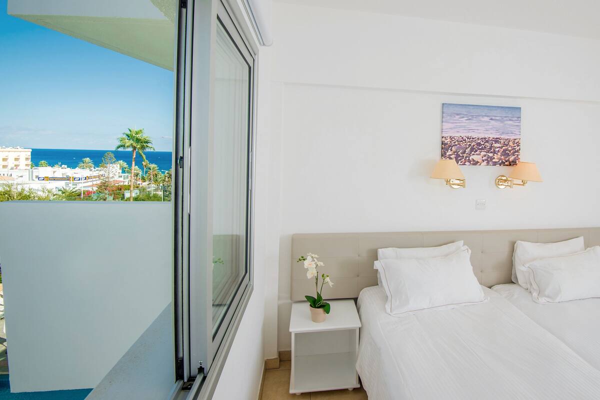A hotel room with a bed, bedside lamps, a nightstand, a potted plant, and a view of the sea and buildings.