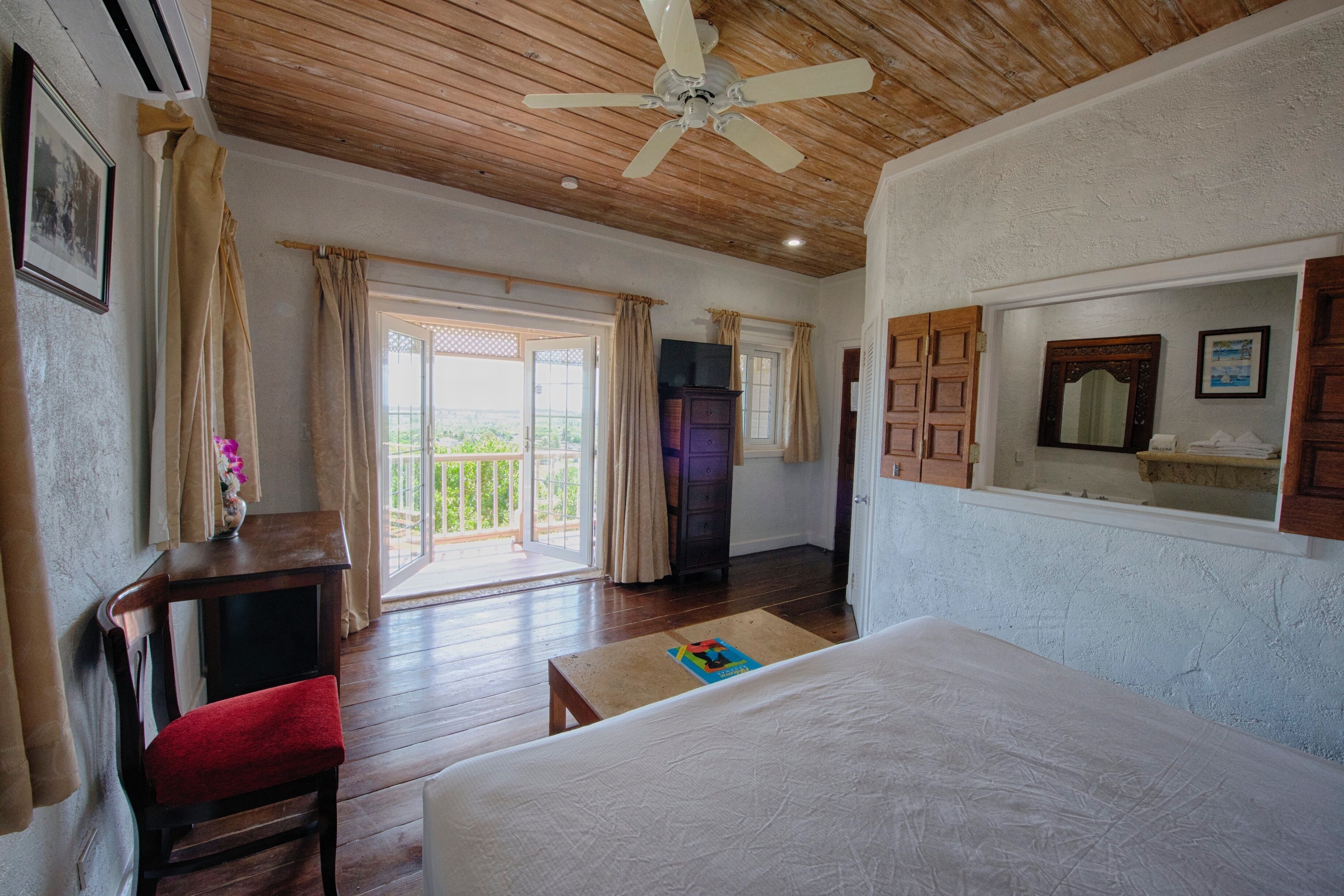 Une chambre avec un lit, un bureau, une chaise, un ventilateur de plafond et une vue sur l’extérieur.