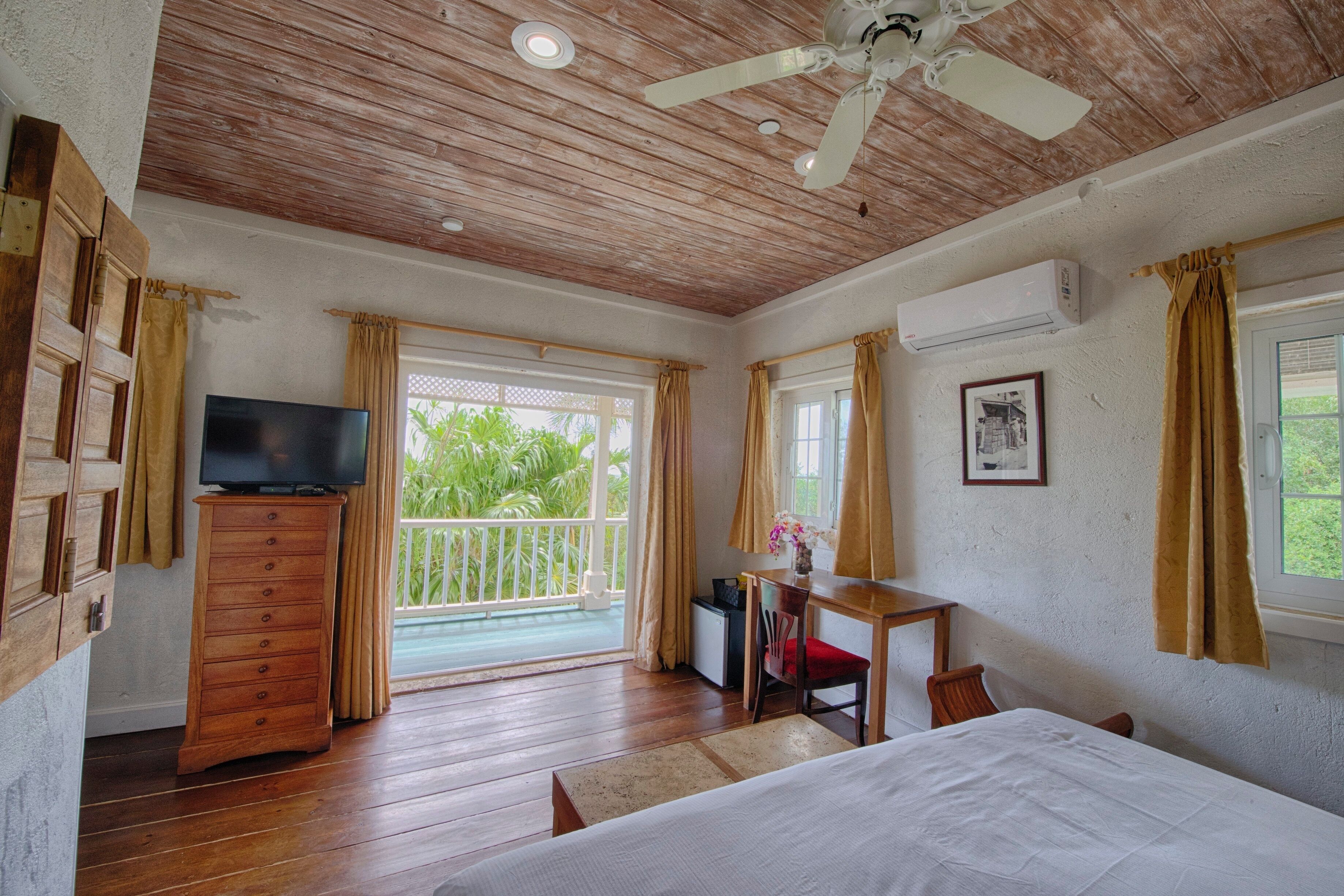 A room with a wooden ceiling, a bed, a TV on a wooden cabinet, a desk with a chair, and a window with curtains.