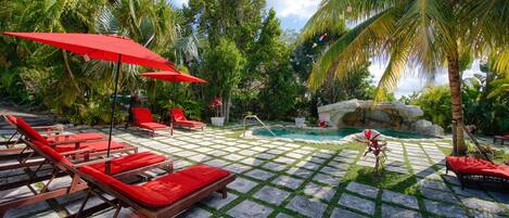 A pool area with lounge chairs and umbrellas, surrounded by palm trees and a stone pathway.