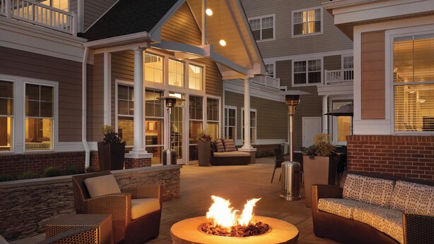 An outdoor patio area with a stone fire pit surrounded by wicker furniture, a brick wall, and a covered seating area with a view of the house. - Residence Inn by Marriott Newport/Middletown (Middletown)