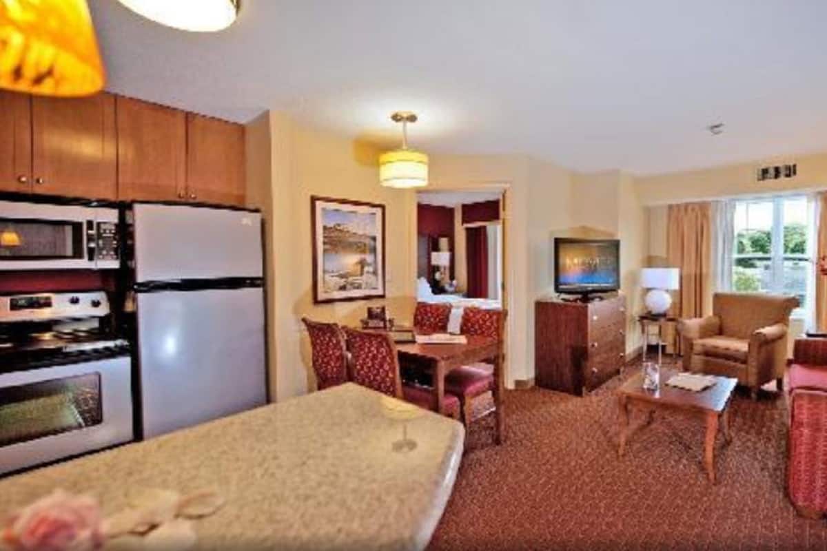 A hotel room with a kitchenette, a dining area, a living room with a sofa and armchairs, a TV, and a window with curtains.