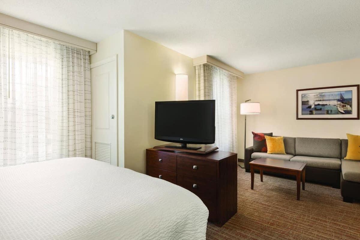 A hotel room with a large bed, a flat-screen TV on a wooden dresser, a sofa with cushions, and a framed picture on the wall.