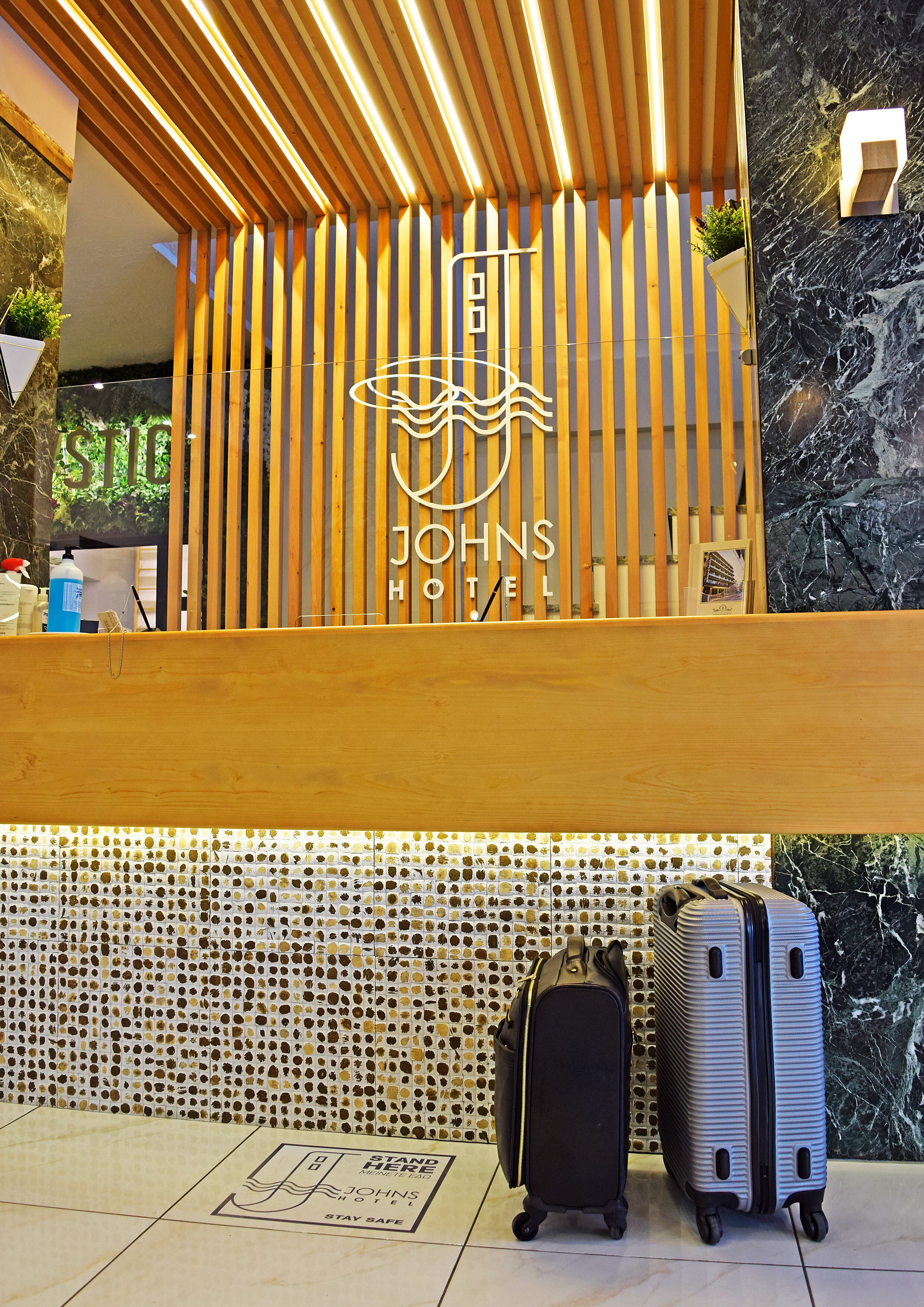 Un comptoir d'accueil à l'hôtel Johns, doté d'un design avec des lattes en bois, d'un logo et de deux valises posées devant.